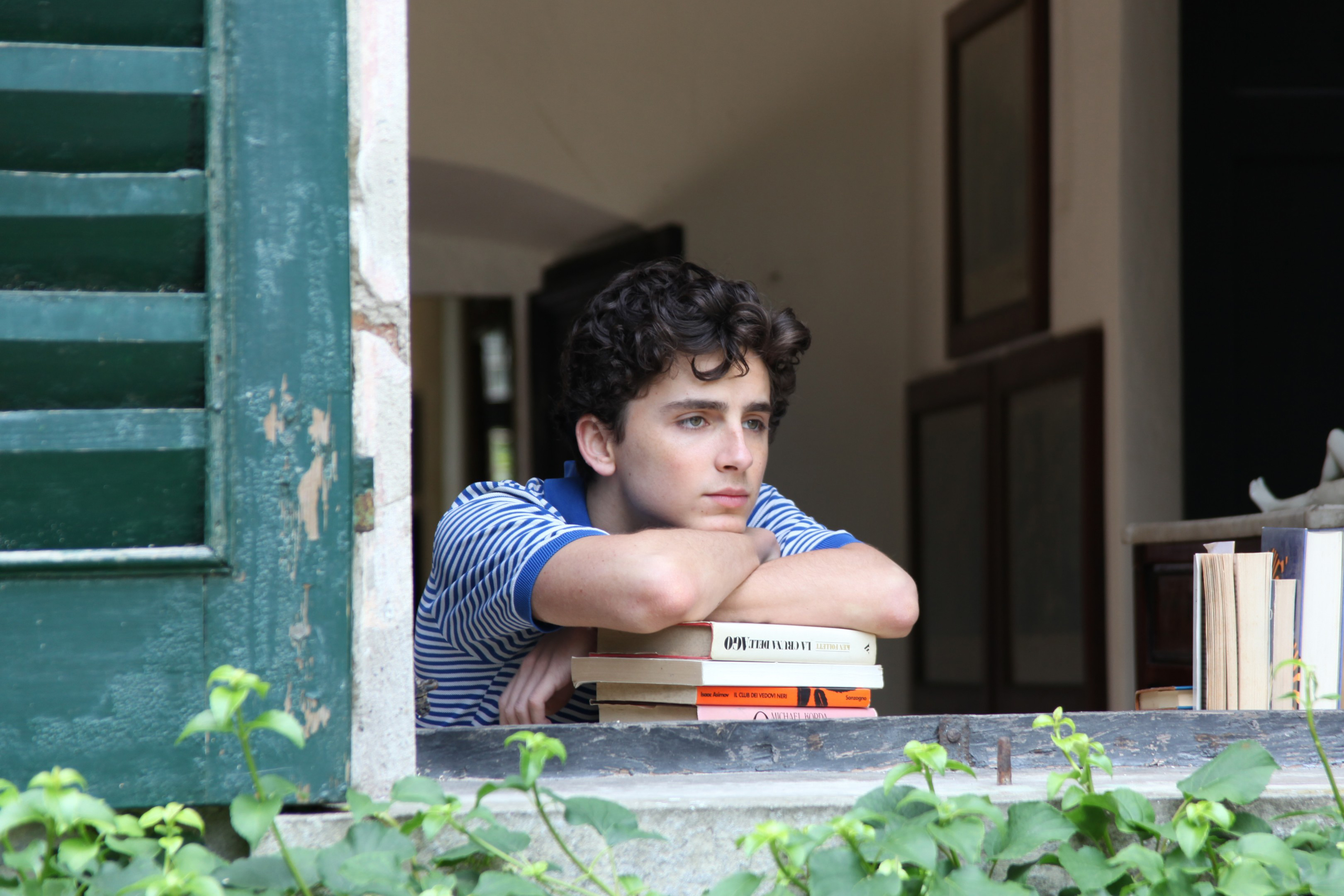 A teenage boy leaning over a pile of books, watching outside